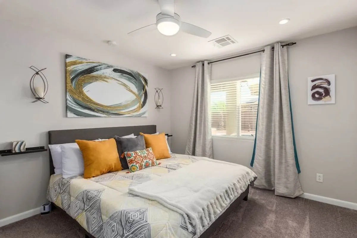 A tranquil bedroom with a bed dressed in colorful pillows and abstract wall art, illuminated by a window streaming in natural light. The space feels peaceful and inviting, with decor touches that evoke Southwestern style.