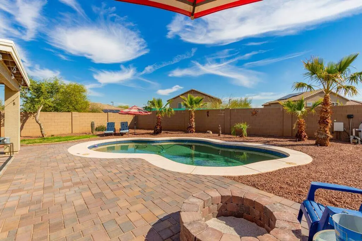 A vibrant backyard oasis featuring a swimming pool surrounded by palm trees, a patio area with umbrella, and a cozy fire pit. The space is set for socializing, relaxing, or gathering after a day out at events.