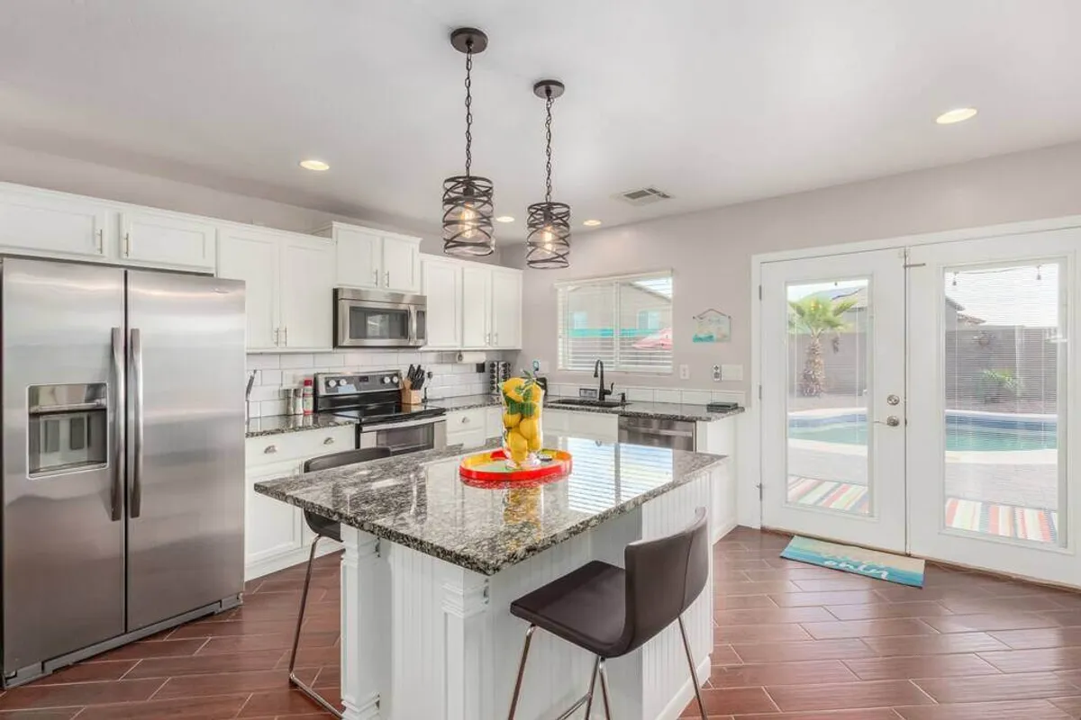 The image focuses on a modern kitchen with a granite countertop, stainless steel appliances, a kitchen island with bar seating, and a decorative fruit centerpiece. The space is bright and designed for both cooking and socializing, seamlessly connecting to the rest of the home.