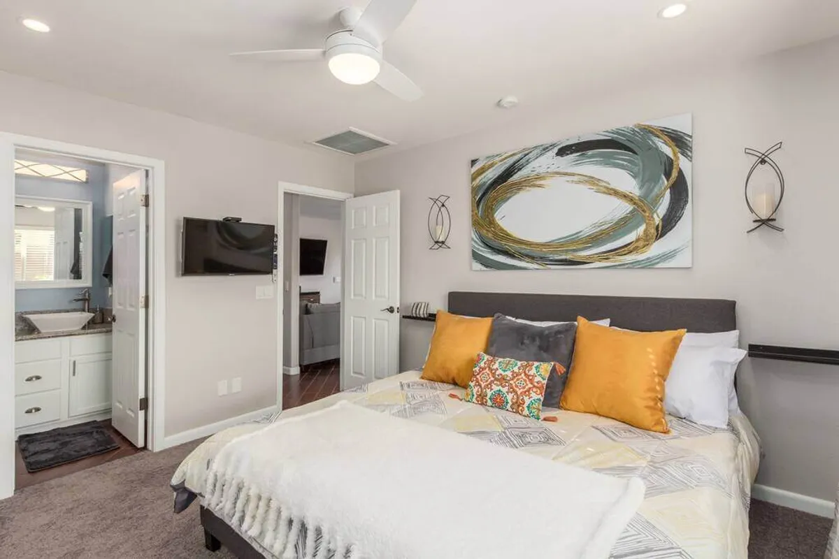 A cozy bedroom featuring a comfortable bed adorned with decorative pillows, abstract Southwestern-inspired wall art, and a door leading to an en-suite bathroom. Soft natural light brightens the restful space, emphasizing relaxation and comfort.