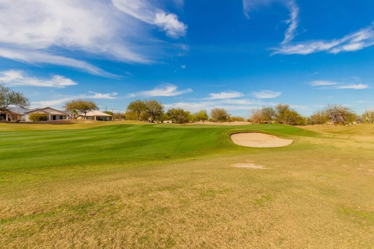 A serene expanse of lush, green grass with a prominent sand bunker under a bright blue sky. Distant homes are visible, hinting at the close proximity between residential comfort and the golf course. The relaxed scene suggests leisure and the tranquility of time spent outdoors.