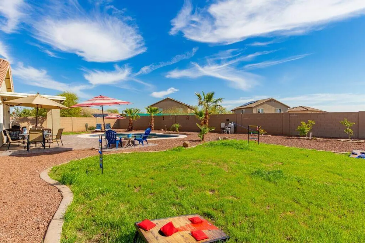 This image features a sparkling pool surrounded by patio seating, outdoor umbrellas, a well-maintained lawn area, and towering palm trees. The scene is set for both relaxation and lively gatherings, capturing people enjoying outdoor dining and lounging by the pool.