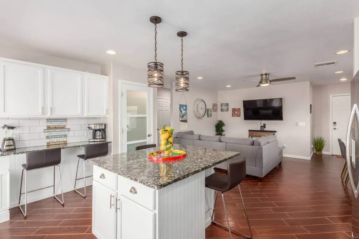 A view from the kitchen area looking toward the living room with a granite kitchen island, bar stools, a cozy sofa, and a wall-mounted TV. The open sightlines highlight the integrated flow between the kitchen and living area, making it ideal for entertaining and family activities.