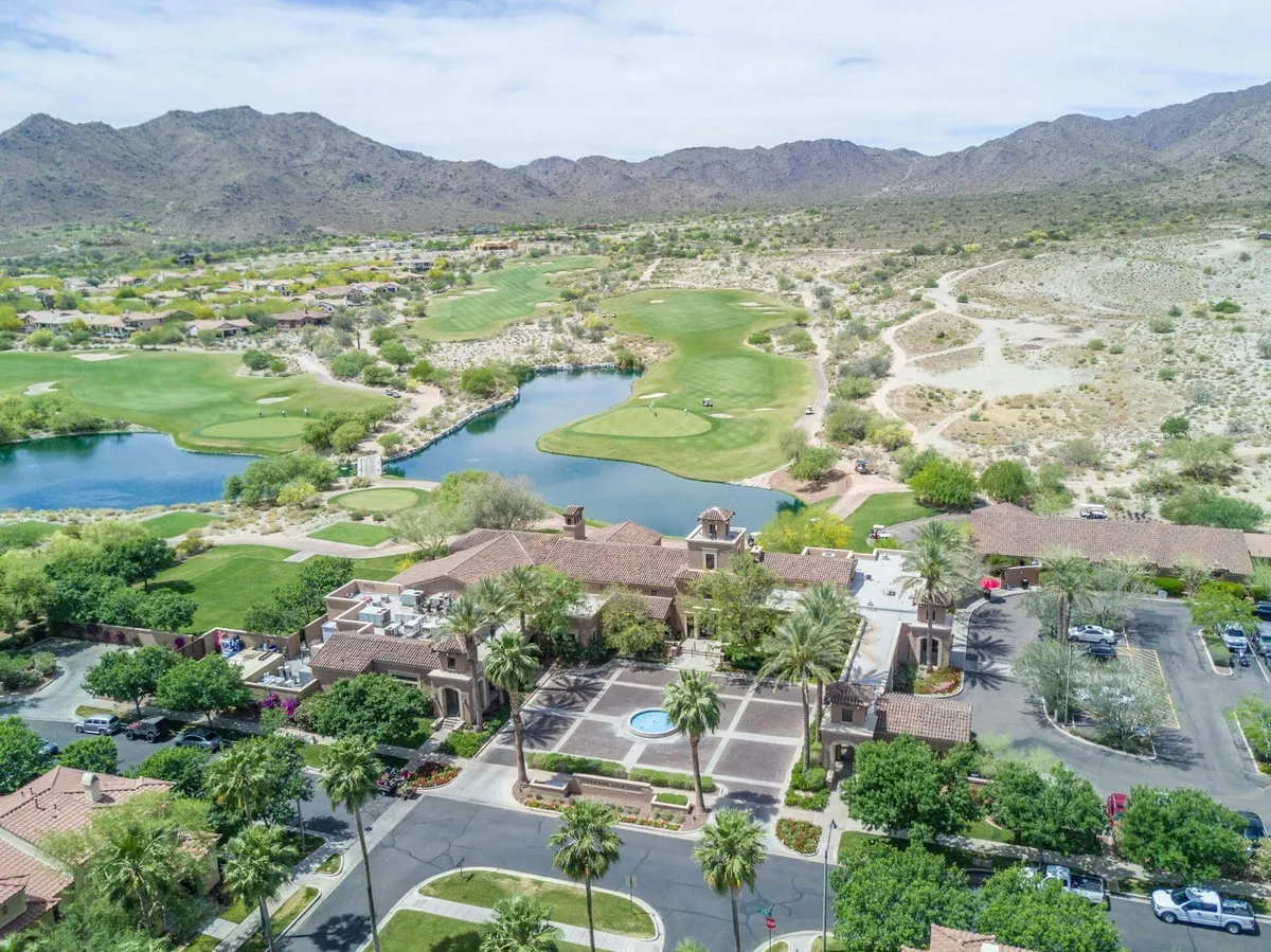 A sweeping view of a golf course bordered by a large residential building, a tranquil lake, and distant mountains. The green is lush and well-maintained, with the early morning or late evening sun casting a soft glow over the landscape, creating a relaxed and upscale atmosphere.
